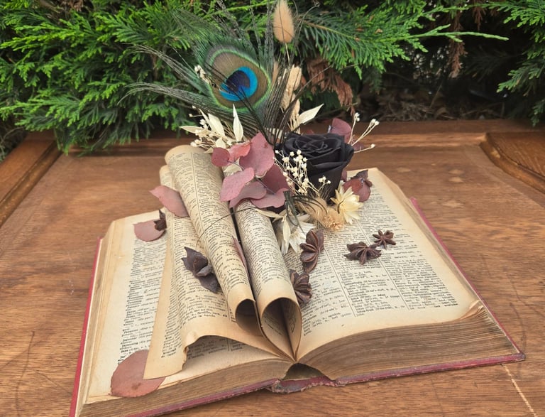 Livre posé avec rose éternelle noire, plume de paon et fleurs séchées, tons pourpre,  réalisé par l'Ensorce'Fleurs