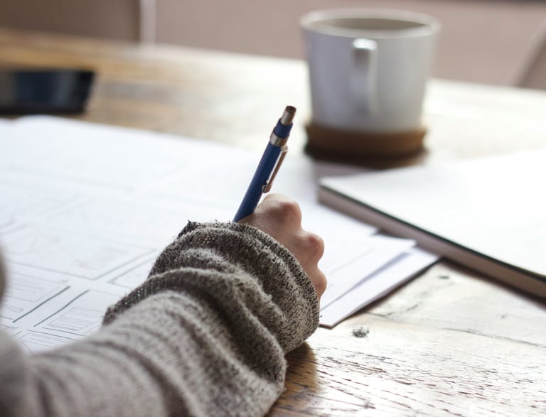 A woman sits at a wooden desk, writing with pen and paper.