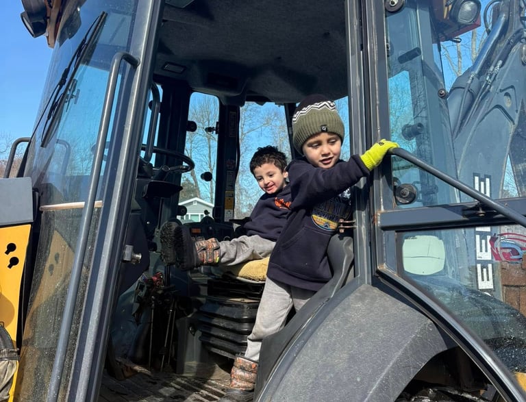 Two young boys playing inside the driver cab of a yellow John Deere construction excavator.
