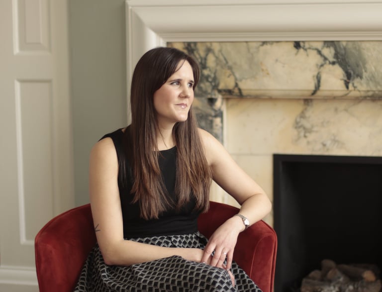 Photo of Anna Joyce expert insomnia therapist sitting in chair by marble fireplace wearing a dress
