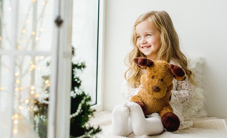 une petite fille qui regarde par la fenetre assise sur un canapé avec un ours en peluche sapin