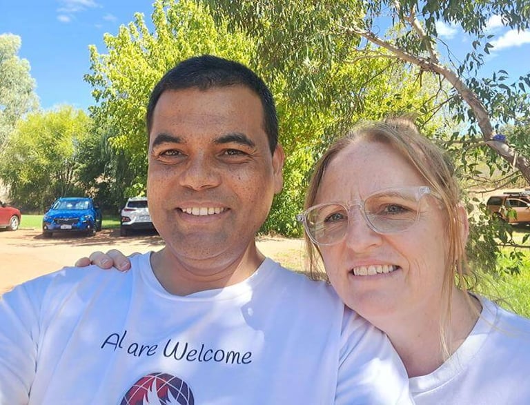 Richard and Salome smiling outdoors, wearing church welcome shirts during a community gathering.