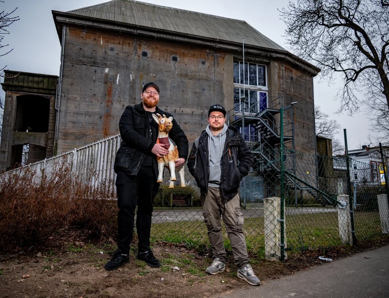 Two men standing outside an industrial bunker building in Hamburg holding a decorative goat statue.