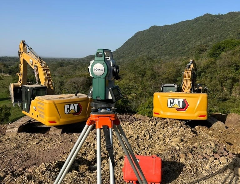 Sokkia total station on a tripod at a construction site with two CAT excavators and hills.