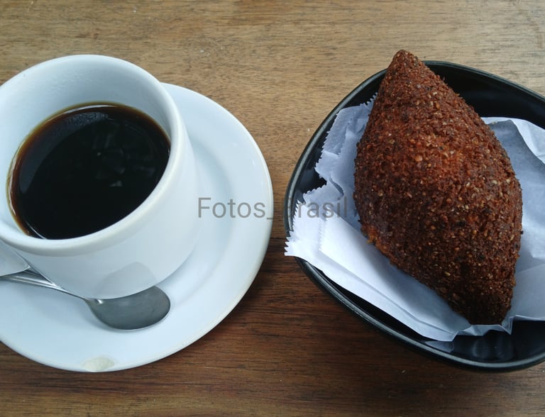 Uma xícara de café fumegante sobre um pires branco, ao lado num pratinho, um kibe dourado e crocante