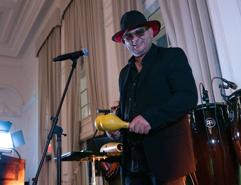 A male musician in a black suit and hat plays yellow maracas with conga drums at a live event.