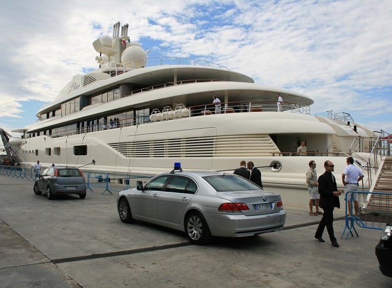 Security vehicle with flashing lights near a 130-meter luxury yacht in Italy, ensuring VIP safety.