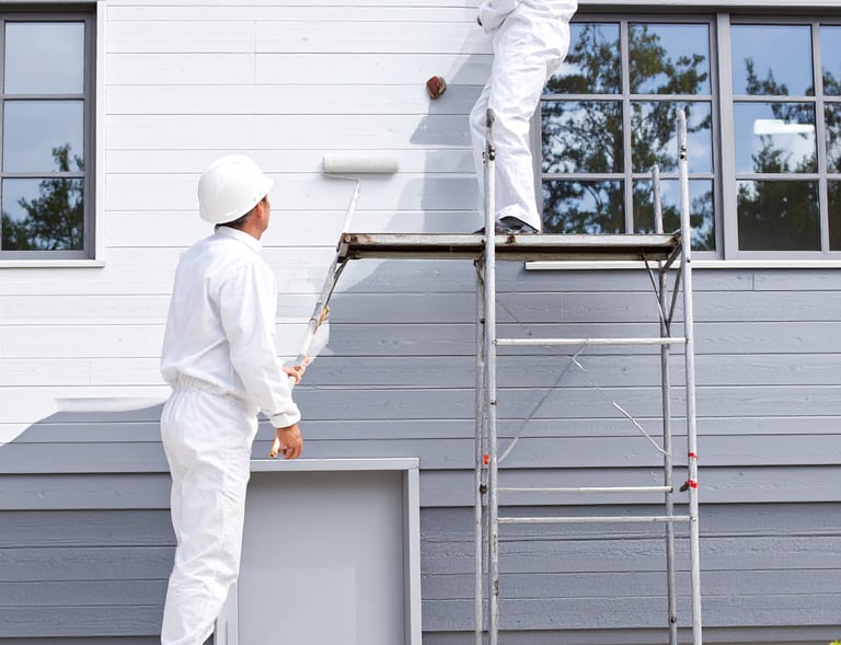 Professional house painters in white coveralls using a roller and scaffolding to paint a home exterior.