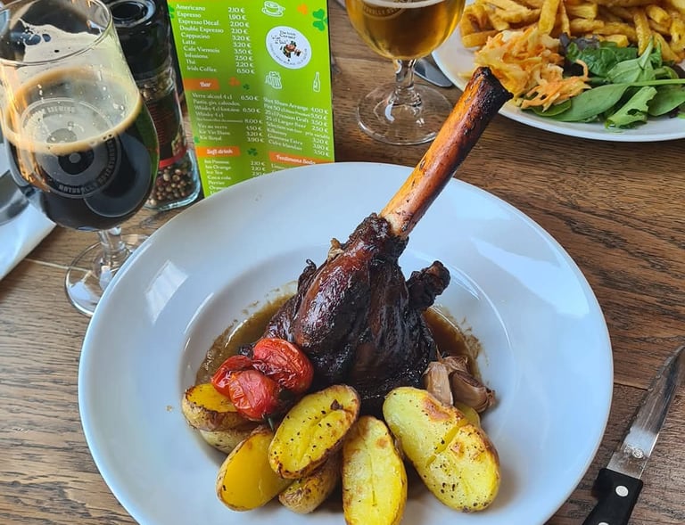 Braised lamb shank served with roasted potatoes, cherry tomatoes, and craft beer at a restaurant table.