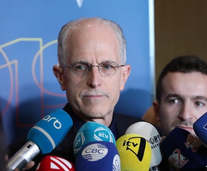 Randy Bennett speaking to reporters surrounded by microphones at a press 