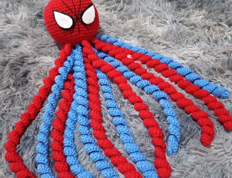 Handmade Spider-Man crochet octopus plush with red and blue curly tentacles on a grey shag rug.