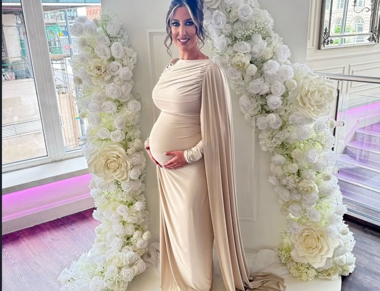 A pregnant woman in a beige maternity gown poses by a white rose flower arch at a baby shower.