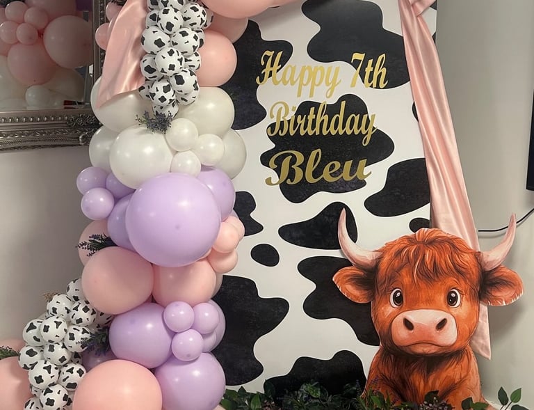 Festive birthday decorations with a cowhide pattern, pink and purple balloons, lush greenery, and a cutout of a cute