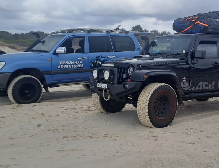 Black Jeep Wrangler and blue Toyota Land Cruiser 4x4 vehicles driving on a sandy beach for off-road adventures.
