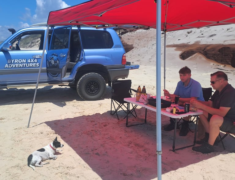 A blue Byron 4x4 Adventures truck parked on a beach with people dining under a red canopy tent.