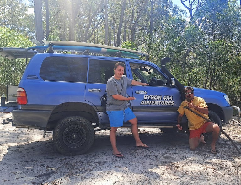 Two men posing next to a blue Byron 4x4 Adventures Toyota Land Cruiser with surfboards in a forest.