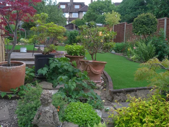 A lush backyard garden featuring terra cotta pots, green lawn, and stone statues for landscaping.