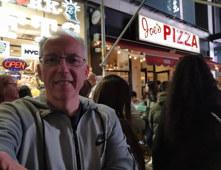 A man in a Nike jacket takes a selfie outside Joe's Pizza in New York City at night.