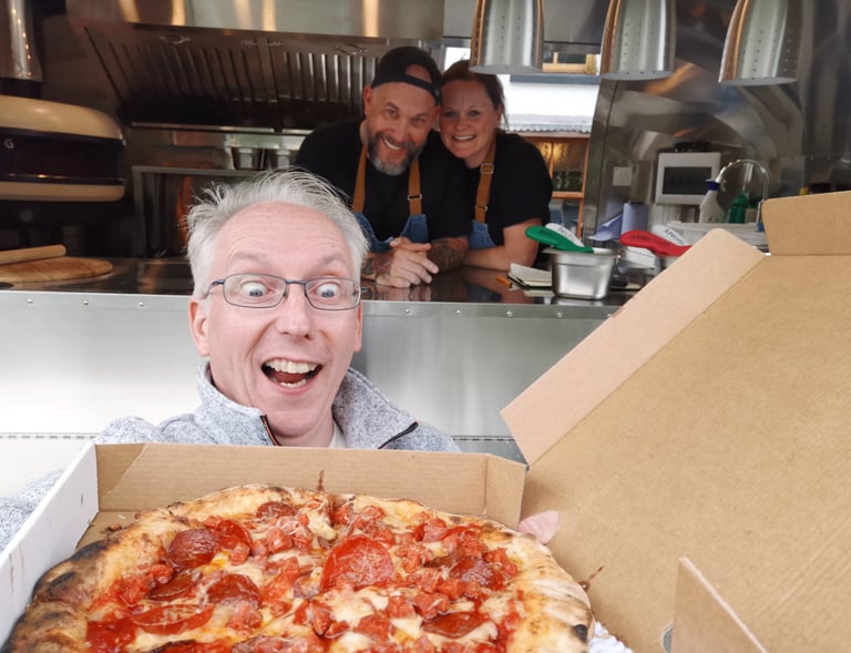 A happy man holds a fresh pepperoni pizza in front of a food truck with two smiling chefs.