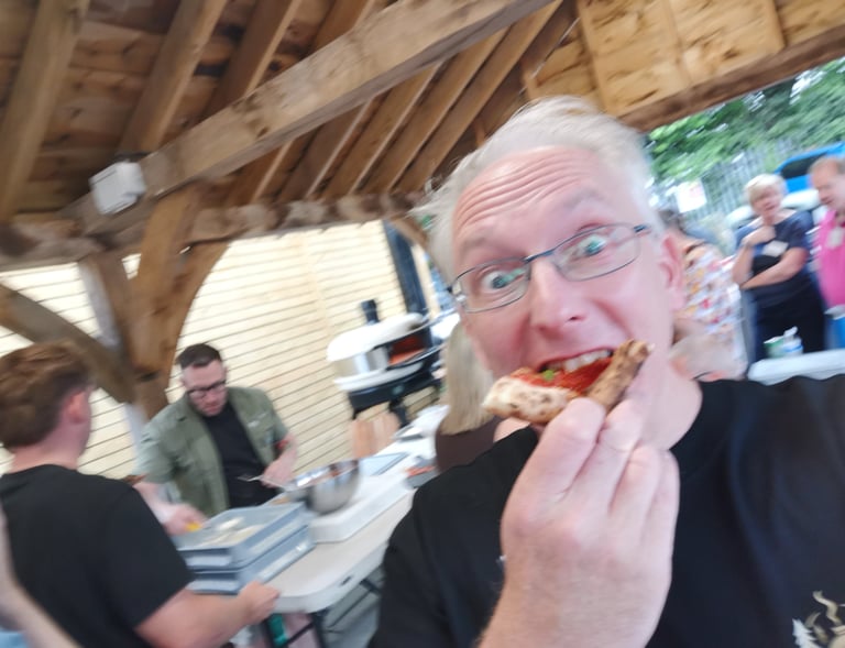 A man wearing glasses eating a slice of wood-fired pizza at an outdoor patio party.