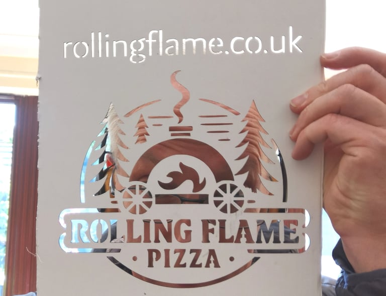 A person holding a laser-cut stencil of the Rolling Flame Pizza logo featuring an outdoor oven and pine trees.