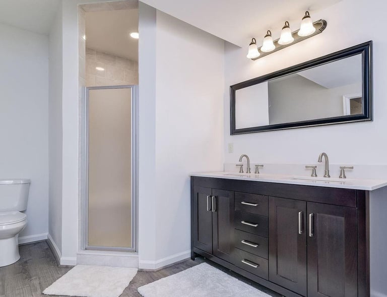 En-suite with custom shower, toilet, and a dual sink vanity. Grey LVT flooring, dark wood vanity.