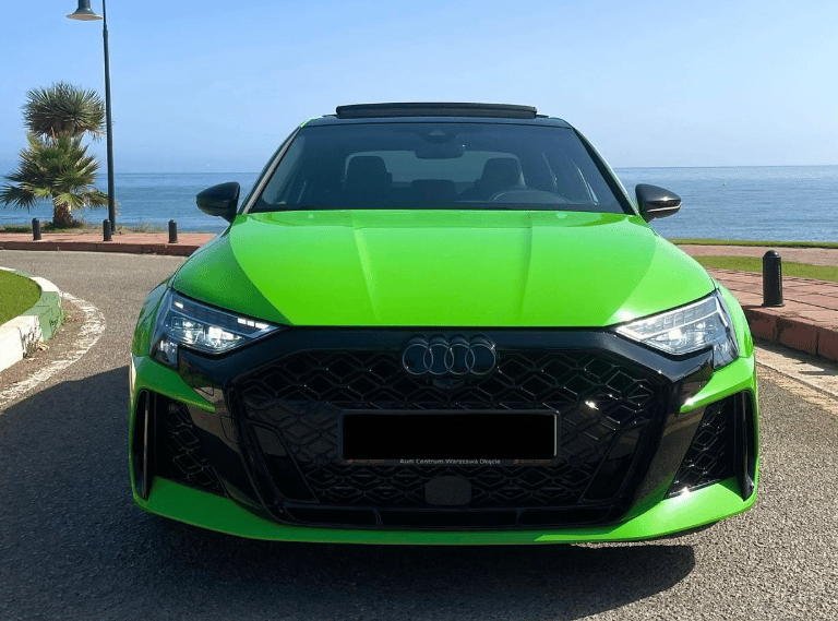 Front view of a bright green Audi RS3 parked on a coastal road with an ocean background.