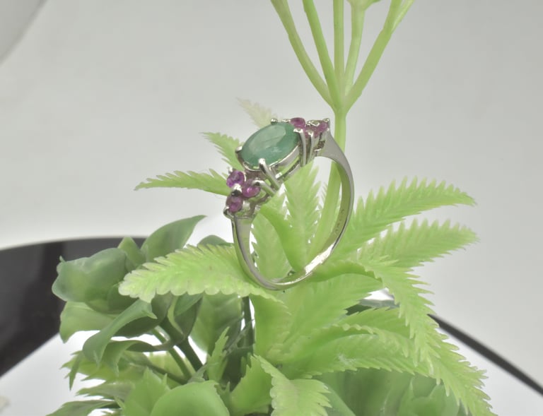 Elegant silver ring featuring an oval green emerald and small ruby side stones on a green plant.