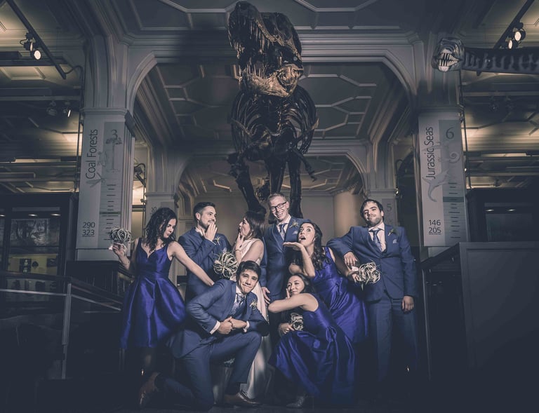 Wedding party in blue attire posing playfully beneath a Tyrannosaurus rex skeleton at a museum venue.