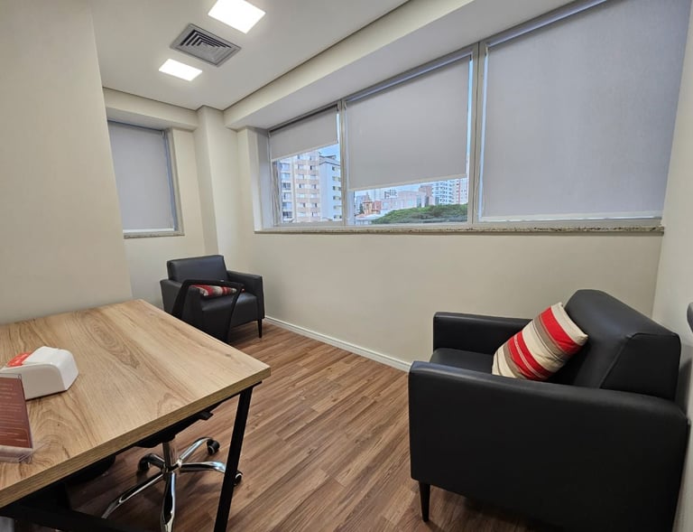 Um consultório de psicologia com duas poltronas de couro preto, uma mesa de madeira clara e uma jane