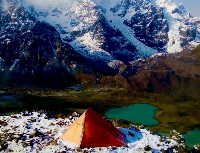 Orange tent at a mountain base camp overlooking snowy peaks and a turquoise alpine lake in Peru.