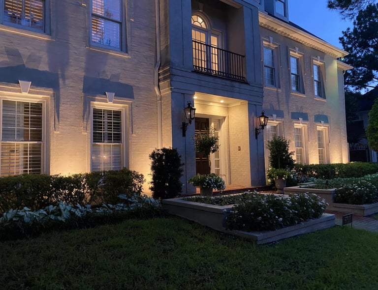 Luxury white brick home at dusk featuring warm exterior landscape lighting and manicured gardens.