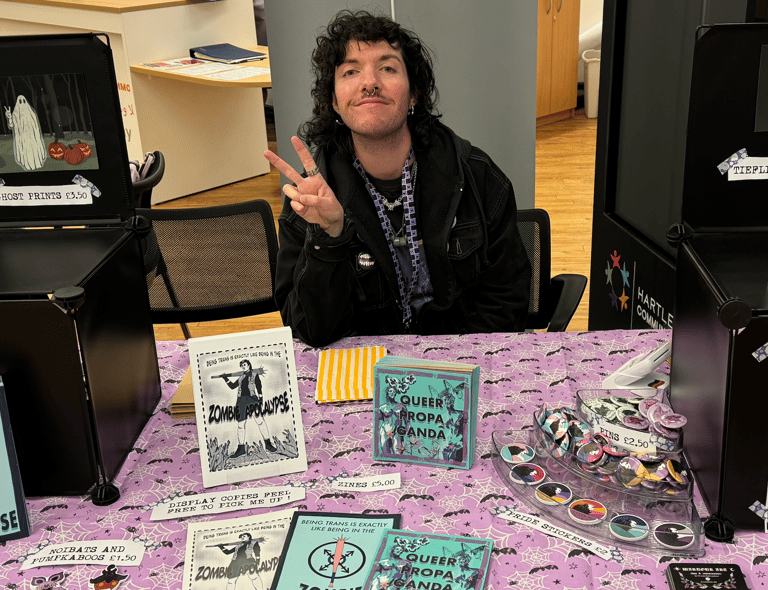 A photograph of the artist, an androgynous person with curly hair, they are doing the peace sign.