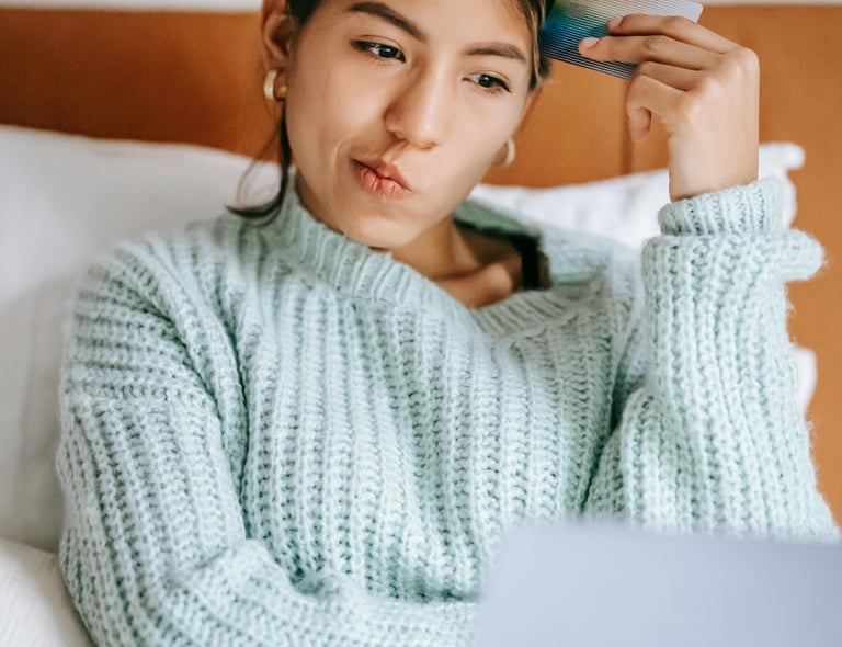 A thoughtful woman holding a credit card while shopping online with a laptop in bed.