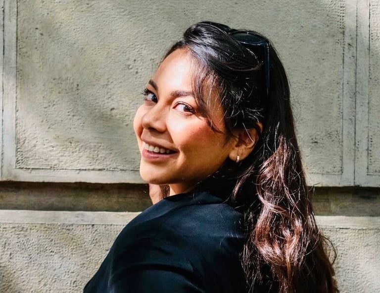 Mujer sonriente de cabello castaño largo con blazer negro frente a un muro de piedra.