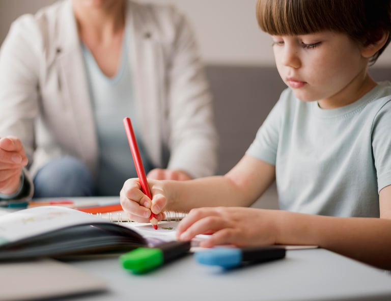Berniukas susikaupęs atlieka mokytojos duodą užduotį, kol mokytoja sėdi šalia.