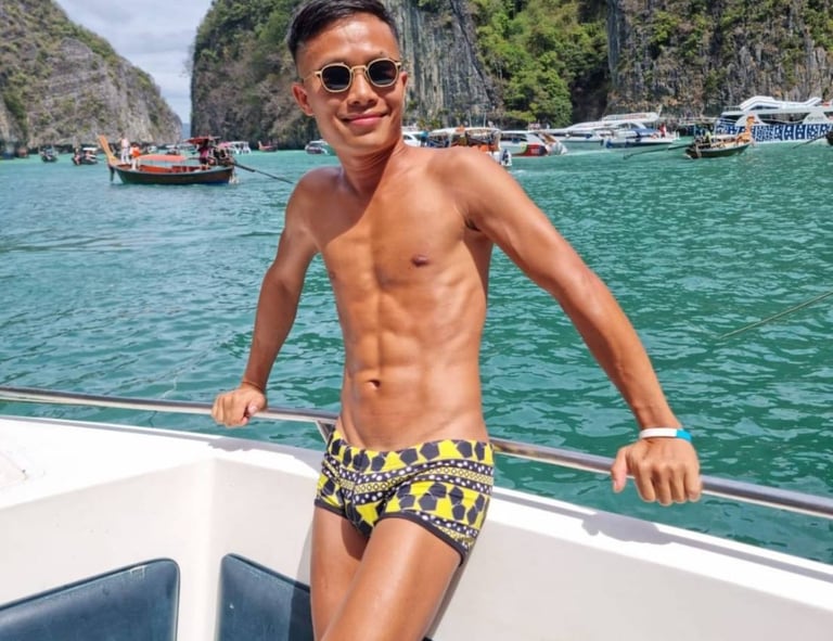 A male outcall masseur in Pattaya standing on a boat wearing swim wear with the sea and an island in the background
