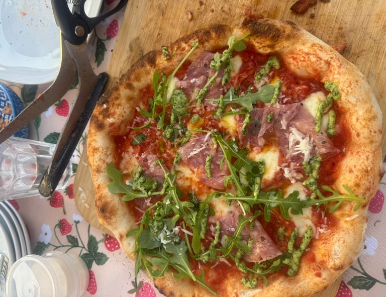 Homemade wood-fired pizza topped with prosciutto, fresh arugula, and pesto drizzle on a wooden board.