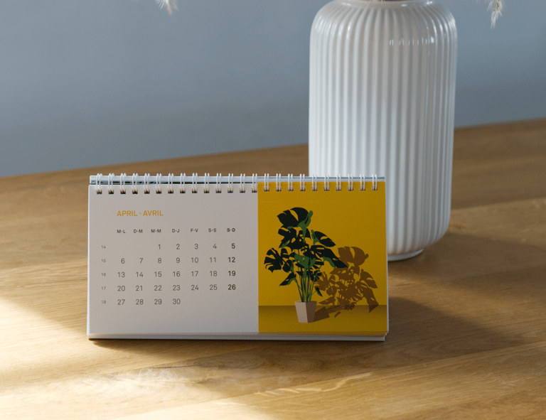Tischkalender für April auf einem Holztisch, mit einer weissen Vase im Hintergrund.