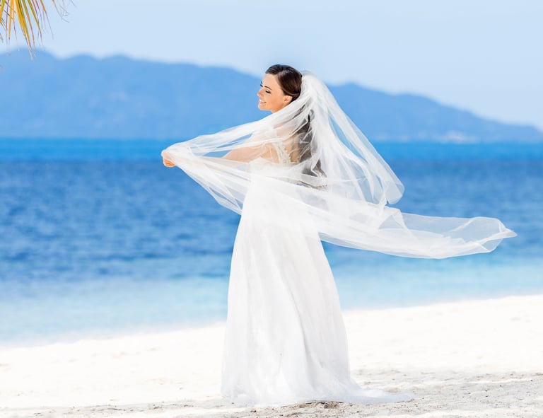 Bride in white dress and veil on sandy Madeira beach with ocean and mountains.