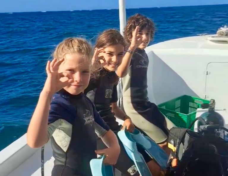 Three children in wetsuits give the OK sign on a dive boat while holding flippers for snorkeling.
