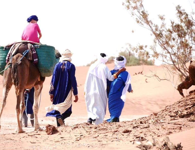 View from behind as three people in nomadic dress walk and one person rides a camel in the Sahara