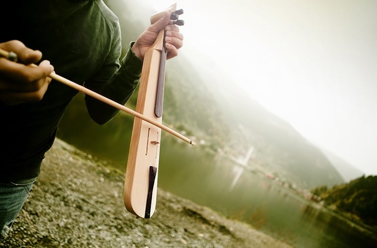 Musician playing a traditional wooden kemence instrument outdoors with a scenic lake and mountain view.