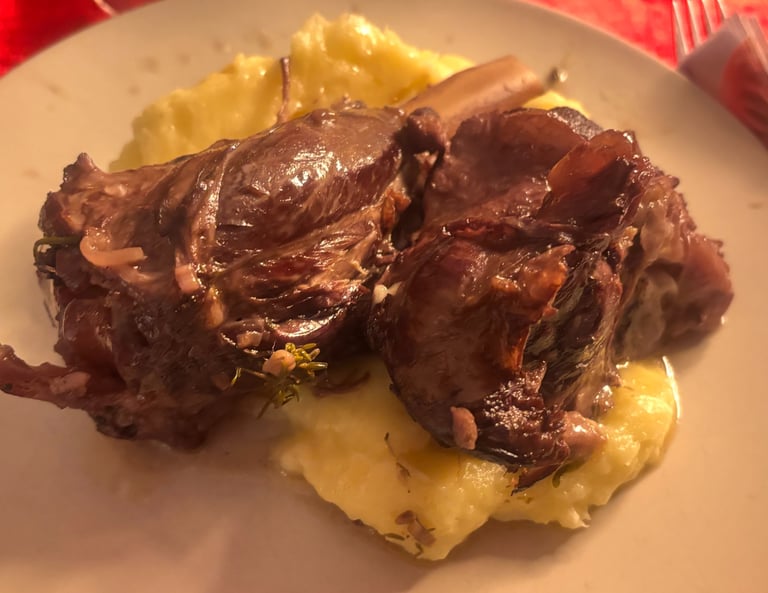 Braised lamb shank served on creamy mashed potatoes on a white plate, set on a red tablecloth.