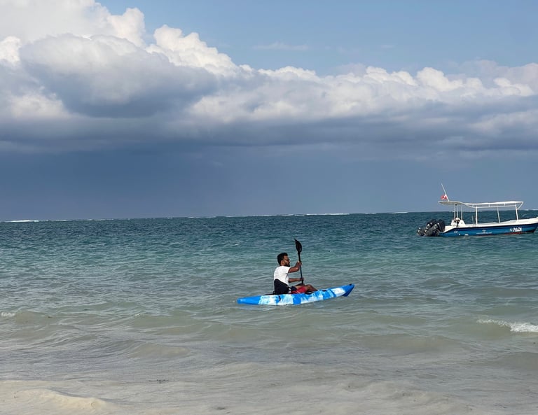 A man paddles a blue camouflage kayak in tropical turquoise ocean waters near a white boat.