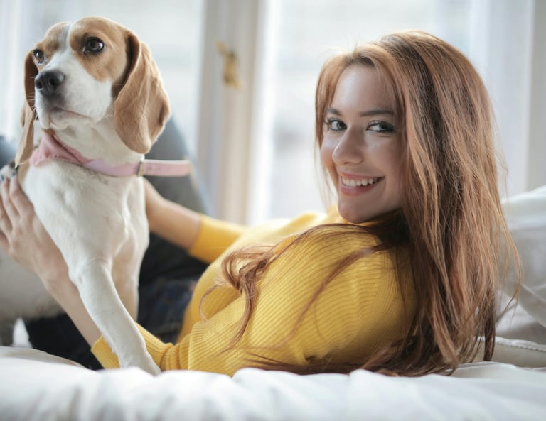 A smiling young woman in a yellow sweater relaxing on a bed with her pet beagle dog.