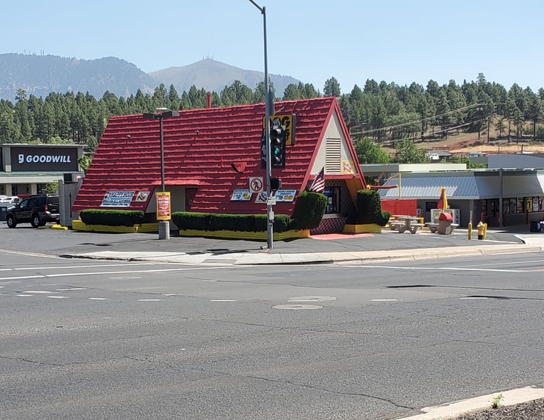 A classic A-frame Wienerschnitzel restaurant with a red roof near a mountain landscape and Goodwill store.