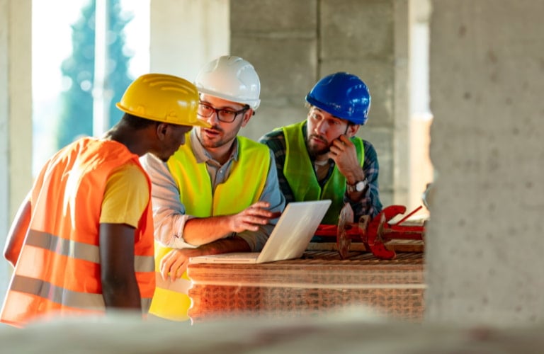Three construction engineers wearing hard hats and safety vests discuss project blueprints on a laptop at a building site.