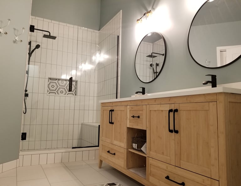 Modern bathroom featuring a light wood vanity, round black mirrors, and a walk-in glass shower with white tile.