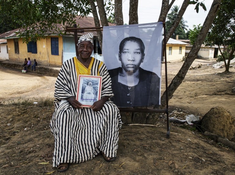Idosa sentado ao ar livre segurando fotografia antiga, ao lado de retrato ampliado em preto